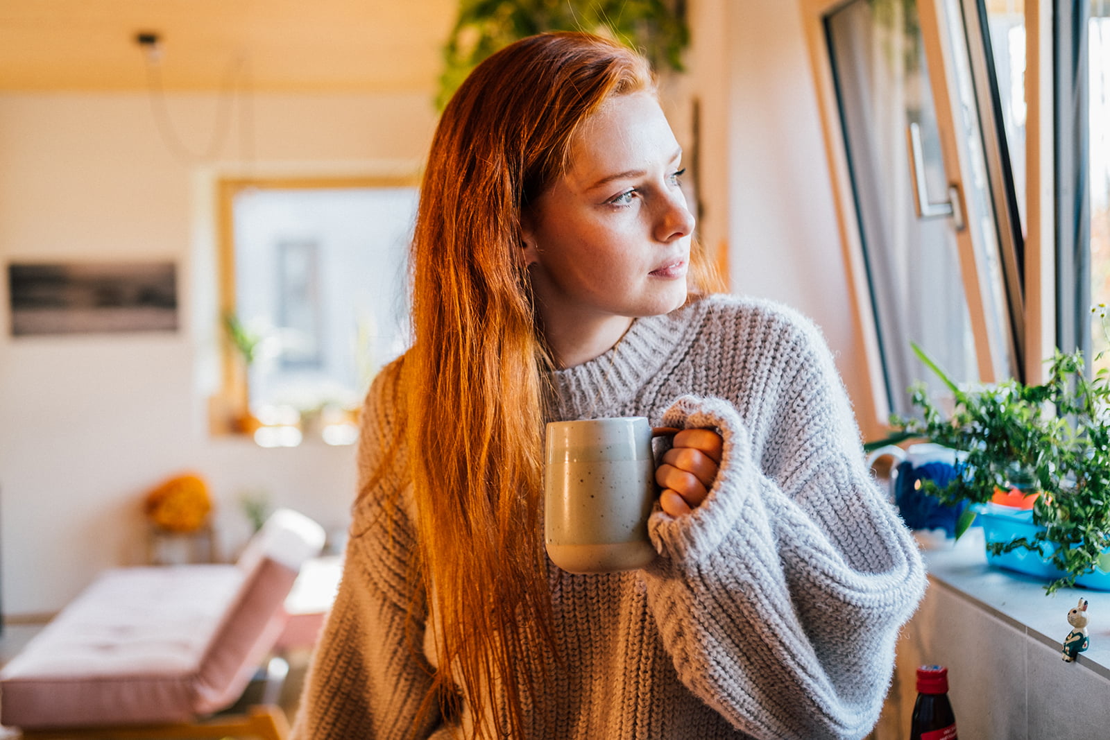 Person som står heime med ein kaffikopp og ser tankefullt ut av vindauget.