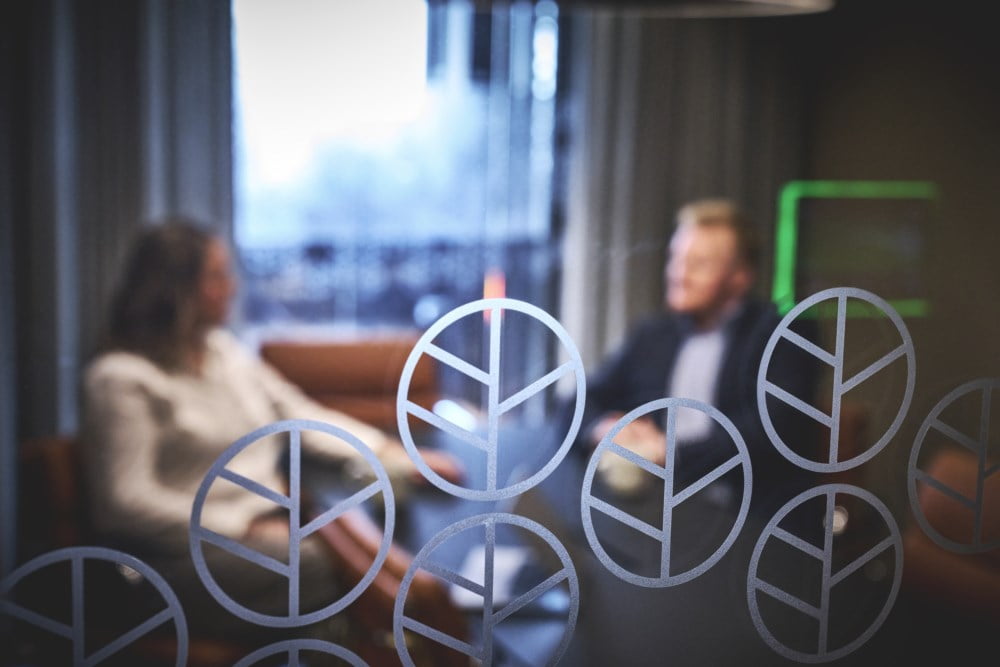 En mann og en kvinne sitter i et møterom og snakker sammen. Vinduet inn mot møterommet reflekterer logoen til Veksthuset