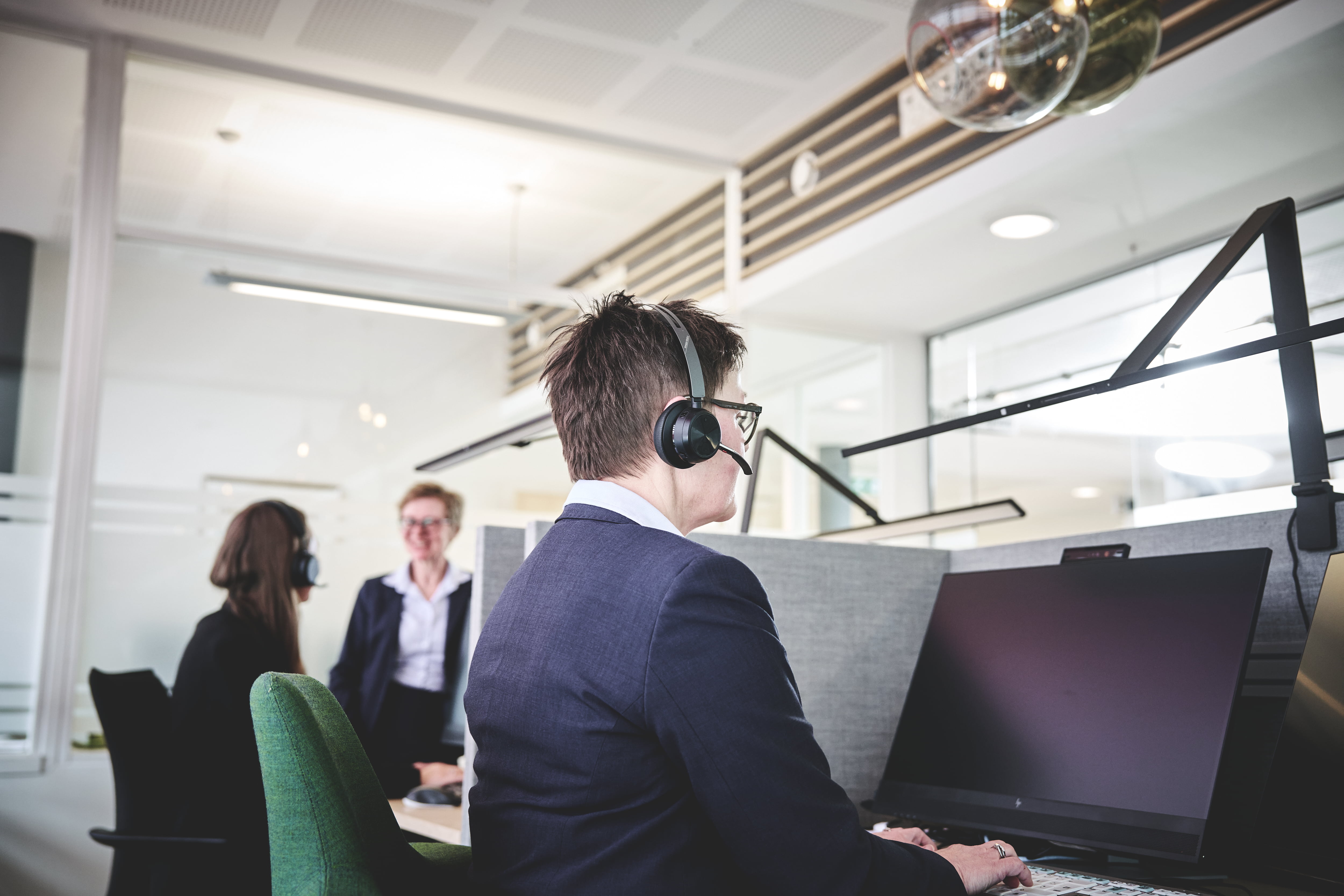 En kvinnelig rådgiver jobber på en pc med hodetelefoner på hodet. I bakgrunnen står to rådgivere og snakker sammen. 